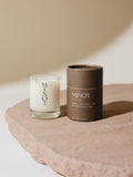 A small white candle in a glass holder labeled "MINOT" sits on a stone surface next to a cylindrical brown cardboard box with text describing the Eventide scent as a blend of amber, bergamot, and sage.