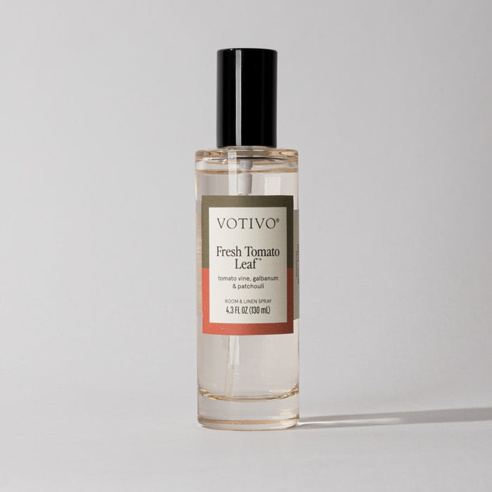 A clear Votivo Fresh Tomato Leaf Room & Linen Spray bottle with a black cap sits on a light background. The label features tomato vine, galbanum, and patchouli fragrance notes and indicates the spray is fabric safe.