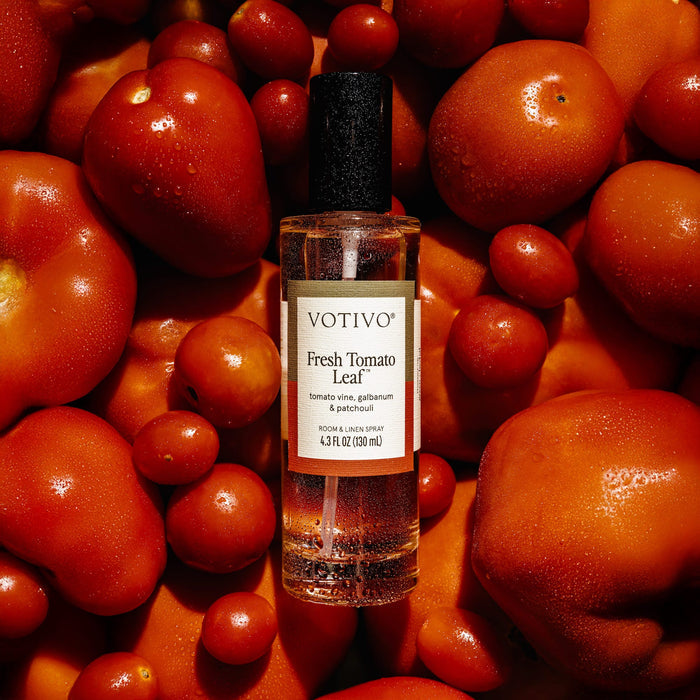 A clear bottle of Votivo Fresh Tomato Leaf Room & Linen Spray with a black cap and white label sits among dewy, ripe red tomatoes, highlighting its fabric-safe and refreshing qualities.