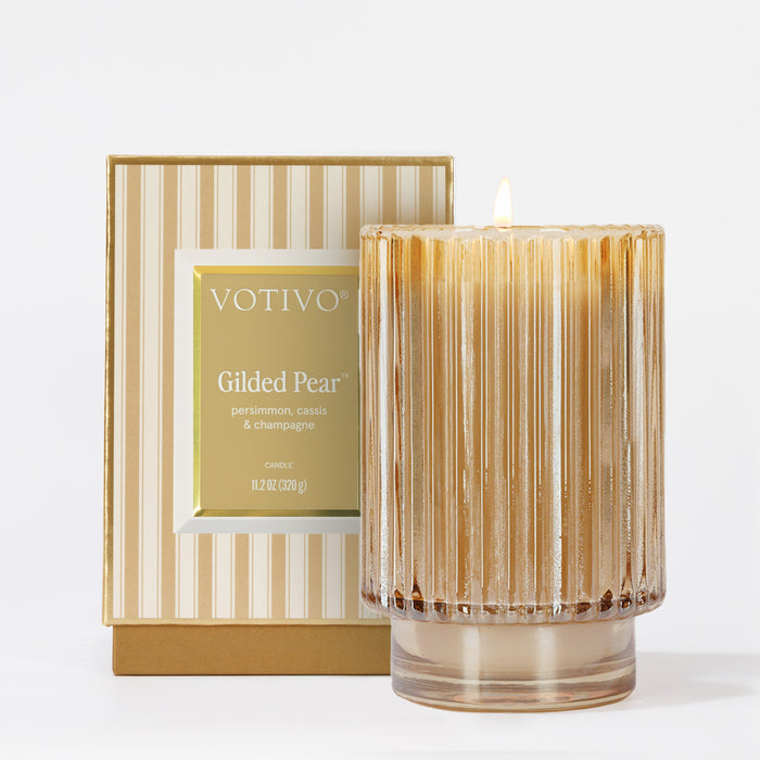 Votivo’s Gilded Pear Holiday Candle glows in a ribbed glass jar next to its matching striped box, featuring scent notes of persimmon, cassis, and champagne.