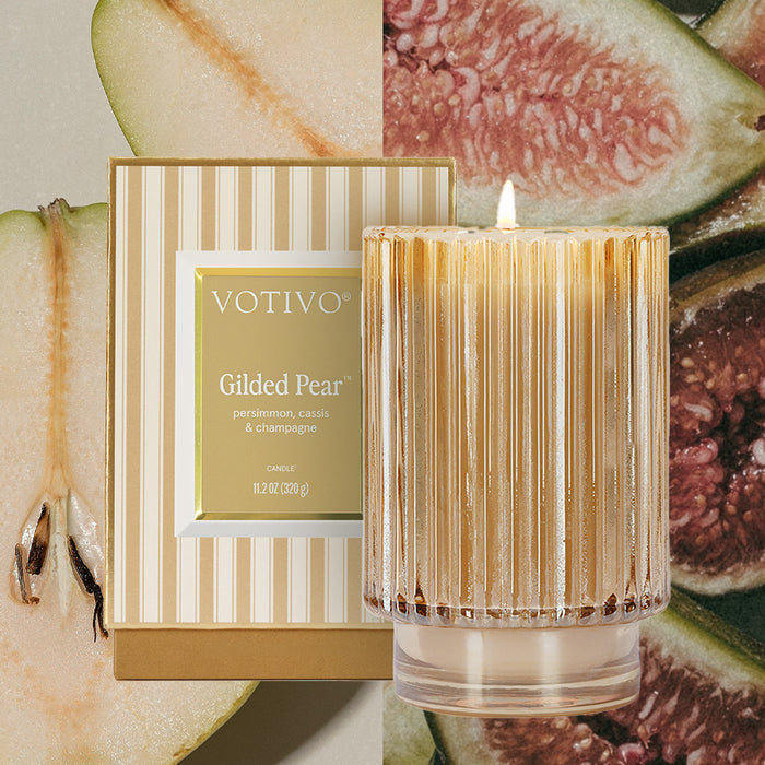 The Votivo Gilded Pear Holiday Candle, crafted from a soy blend and presented in a ribbed glass vessel, is lit beside its gold and cream striped box, with sliced pears and figs adding festive flair in the background.