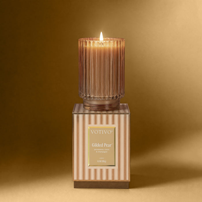 A lit Gilded Pear ribbed candle vessel sitting on a striped box against a gold background