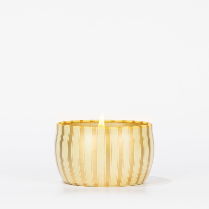 A single lit Votivo Gilded Pear Holiday Tin Candle, in a round gold tin with vertical stripes, is set against a plain white background.