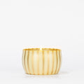 A single lit Votivo Gilded Pear Holiday Tin Candle, in a round gold tin with vertical stripes, is set against a plain white background.