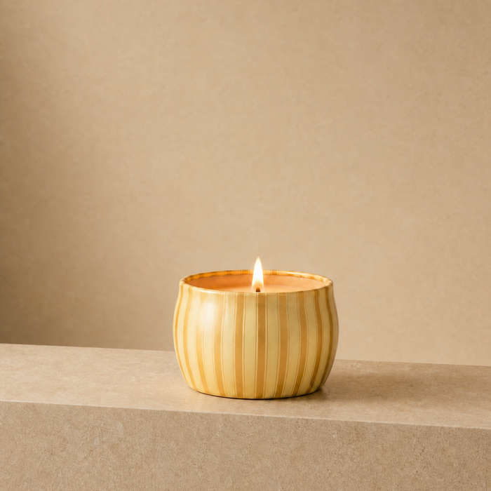A lit Gilded Pear tin candle sits on a matching textured beige surface against a solid beige background.