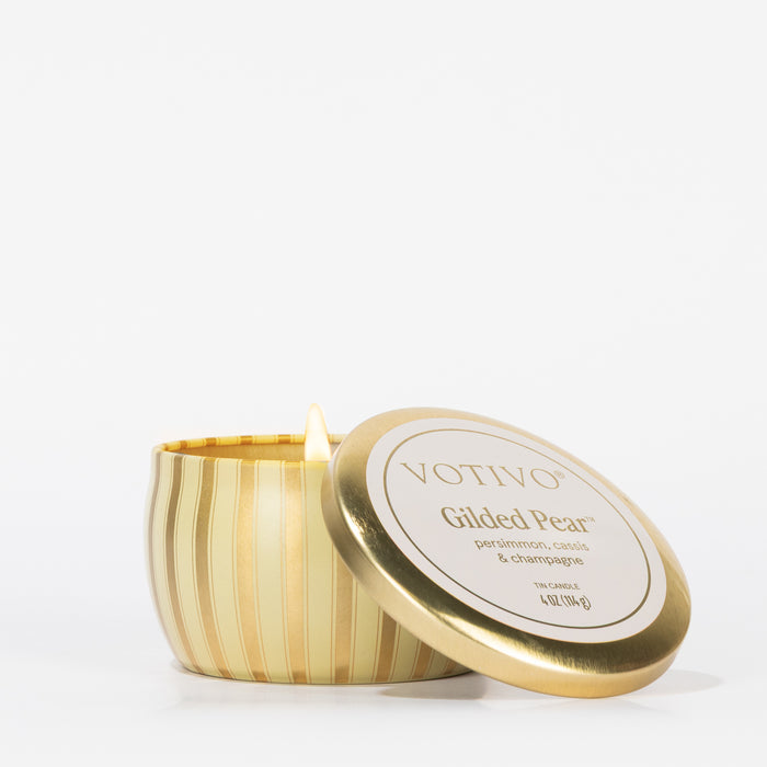 A Votivo Gilded Pear Holiday Tin Candle with gold stripes, made from a soy blend wax and featuring notes of persimmon, cassis, and champagne, sits open with its lid leaning against it on a white background.