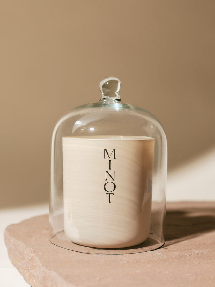 A MINOT candle is encased in a clear glass cloche, placed on a textured stone surface, with a soft, neutral background.