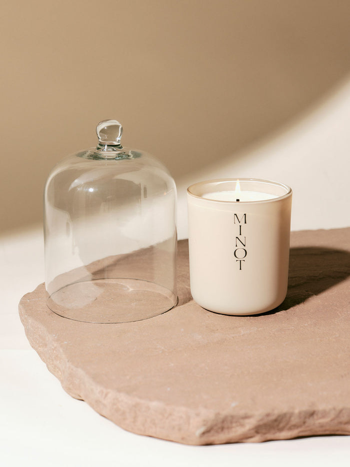 A lit MINOT candle sits on a smooth stone surface next to a glass cloche.