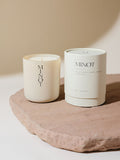 A non-toxic soy candle in a glass vessel labeled "MINOT" sits on a textured stone surface next to a white cylindrical box labeled "Haven: Black Currant, Jasmine, Vanilla"
