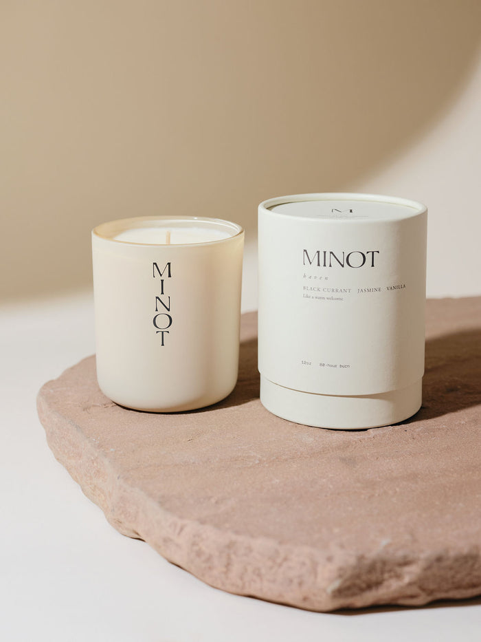 A non-toxic soy candle in a glass vessel labeled "MINOT" sits on a textured stone surface next to a white cylindrical box labeled "Haven: Black Currant, Jasmine, Vanilla"