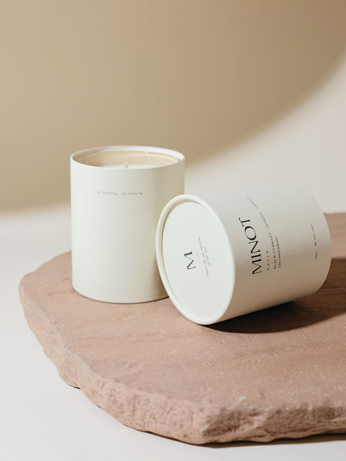White cylindrical packaging holds a MINOT Haven scented candle with its lid tilted to the side on a textured stone slab. The background is a soft beige color.