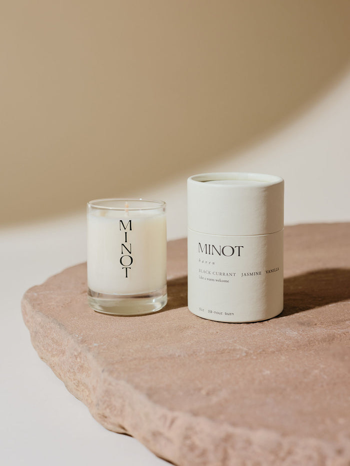 A small soy candle in a glass holder labeled "MINOT" sits on a stone surface next to a cylindrical white cardboard box with text describing the Haven scent as a blend of black currant, jasmine, and vanilla.