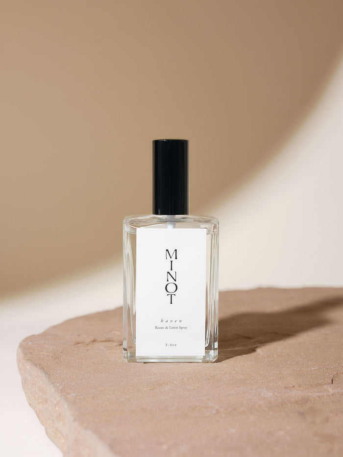 A bottle of MINOT Haven Room & Linen Spray with a black cap, placed on a stone surface. The background is softly lit in beige tones.