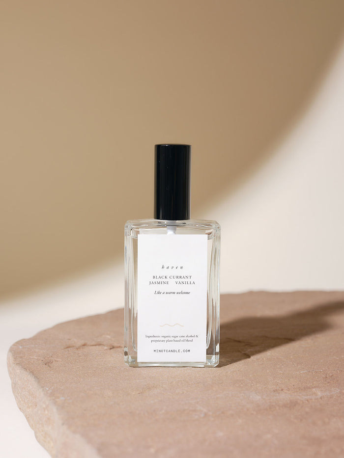 A glass bottle of black currant, jasmine, and vanilla scented room and linen spray with a black cap, placed on a textured stone surface against a soft beige background.