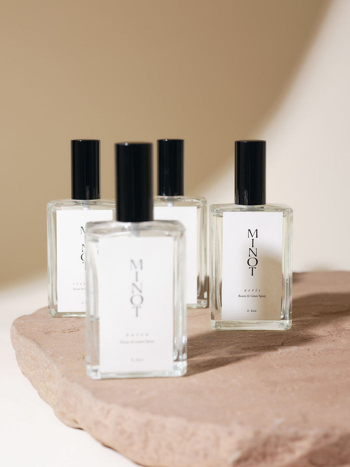 Four glass bottles with black caps labeled "MINOT Room & Linen Spray" sit on a textured stone surface. Soft lighting creates a warm ambiance.