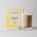 The Votivo Honeysuckle 10 oz. Signature Candle features soy blend wax in a clear glass and comes in a yellow and cream box with bold black text displaying the scent name and product details.