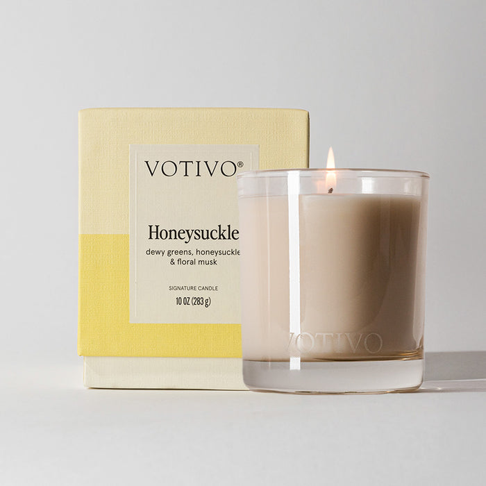 The Votivo Honeysuckle 10 oz. Signature Candle features soy blend wax in a clear glass and comes in a yellow and cream box with bold black text displaying the scent name and product details.