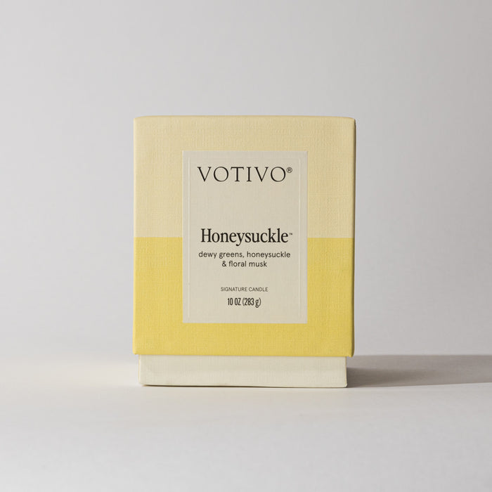 The Votivo Honeysuckle 10 oz. Signature Candle features soy blend wax, dewy greens, honeysuckle, and floral musk notes. It comes in a cream and yellow box with black text on a plain light background.
