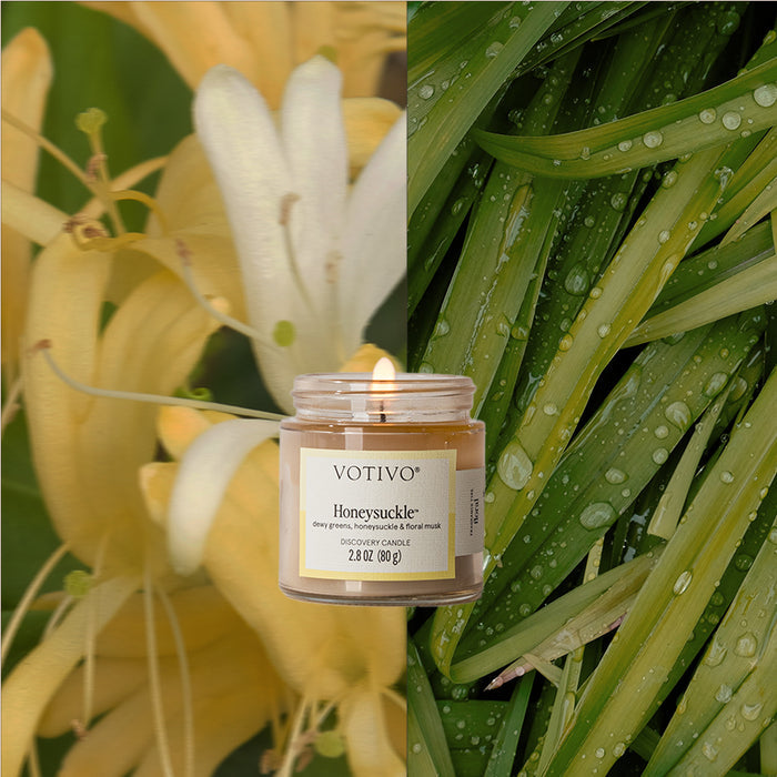 A Votivo Honeysuckle Discovery Candle burns before a split backdrop: close-up yellow honeysuckle blooms on the left and fresh green dewy grass on the right, highlighting the floral fragrance’s essence.