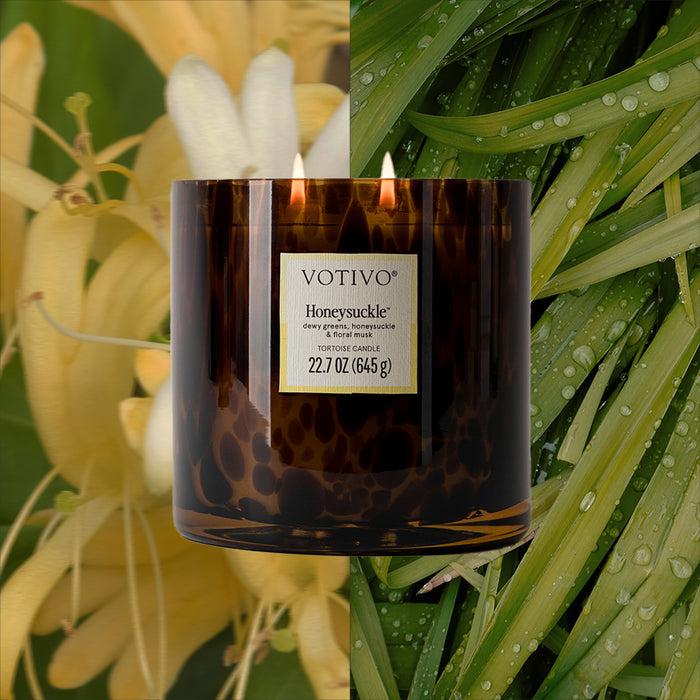 A dark glass Votivo Honeysuckle Tortoise Candle with two flames sits before a background split between yellow honeysuckle flowers and dewy green grass. Made from soy blend wax.