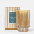 The Votivo Icy Blue Pine Holiday Candle glows in a ribbed glass holder beside its gold and white striped box, filling the air with fresh green, fir needle, and cedar notes from a soy blend wax.