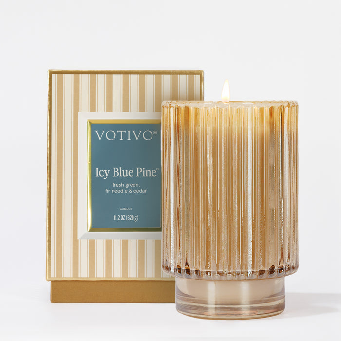 The Votivo Icy Blue Pine Holiday Candle glows in a ribbed glass holder beside its gold and white striped box, filling the air with fresh green, fir needle, and cedar notes from a soy blend wax.