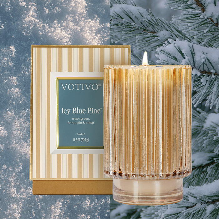 The Votivo Icy Blue Pine Holiday Candle, crafted from a soy blend wax, rests in a ribbed glass container next to its striped box, accented by snow-dusted pine branches in the background.