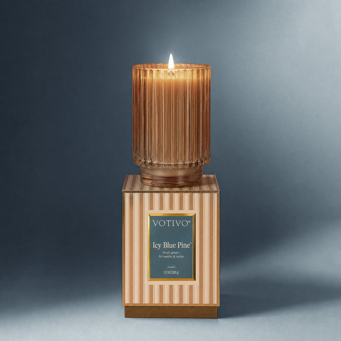 The Votivo Icy Blue Pine Holiday Candle, made with a soy blend wax, glows softly from its ribbed glass holder on a striped box against a gray background.