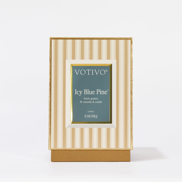 The Votivo Icy Blue Pine Holiday Candle features fresh green, fir needle, and cedar notes in a gold and cream striped box with a green label. This soy blend wax candle stands upright against a white background.