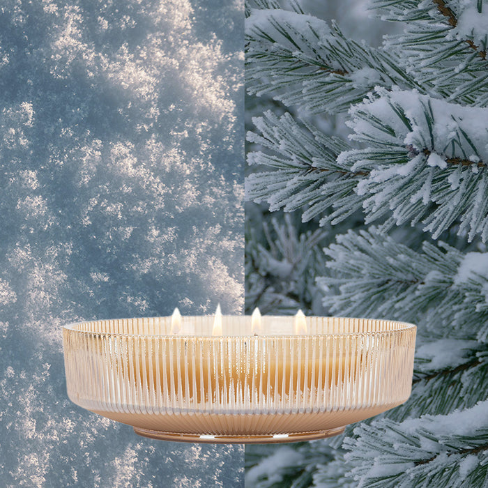 The Votivo Icy Blue Pine Holiday Multi-Wick Candle, crafted with a soy blend and presented in a glass bowl, glows amidst frosty snow on the left and snow-tipped Icy Blue Pine branches on the right.