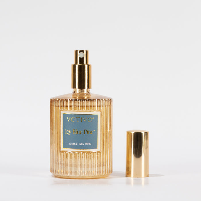 A gold ribbed glass bottle of Votivo Icy Blue Pine Room & Linen Spray with a matching gold cap, set against a white background, delivers a burst of fresh fragrance to any space.