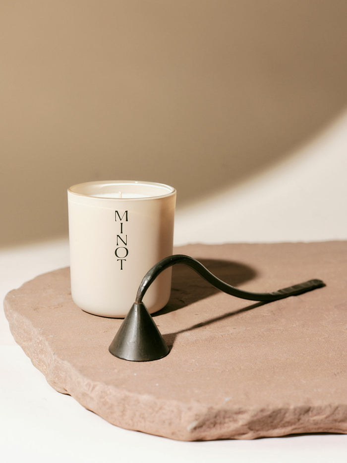 A MINOT candle stands on a stone slab beside a dark metal snuffer, set against a beige background.