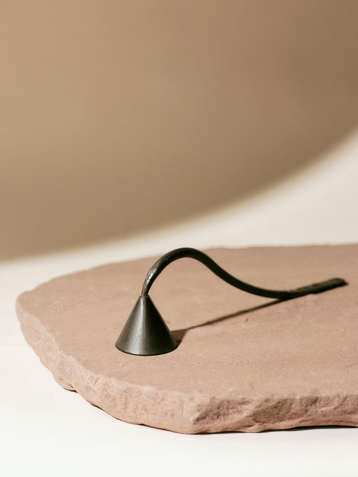 A metal candle snuffer with a curved handle rests on a textured stone slab against a soft beige background.