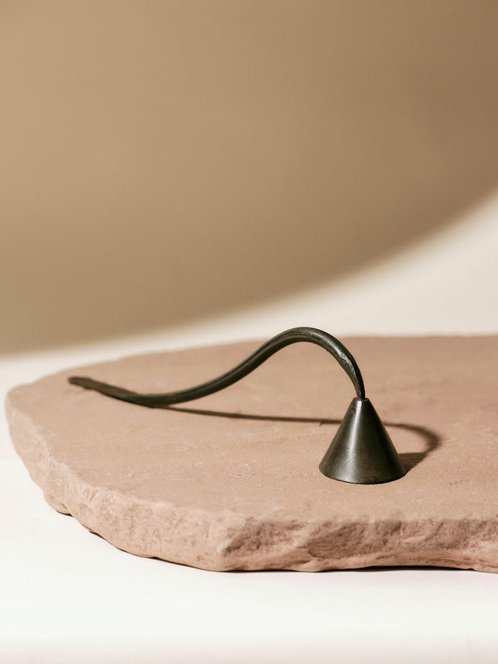 A sleek metal candle snuffer with a curved handle rests on a textured, stone-like surface against a neutral background.