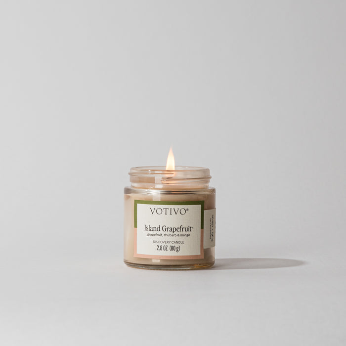 A Votivo Island Grapefruit Discovery Candle glows in a clear glass jar against a white background. Made with soy blend wax, its label features notes of grapefruit, rhubarb, and tangerine.