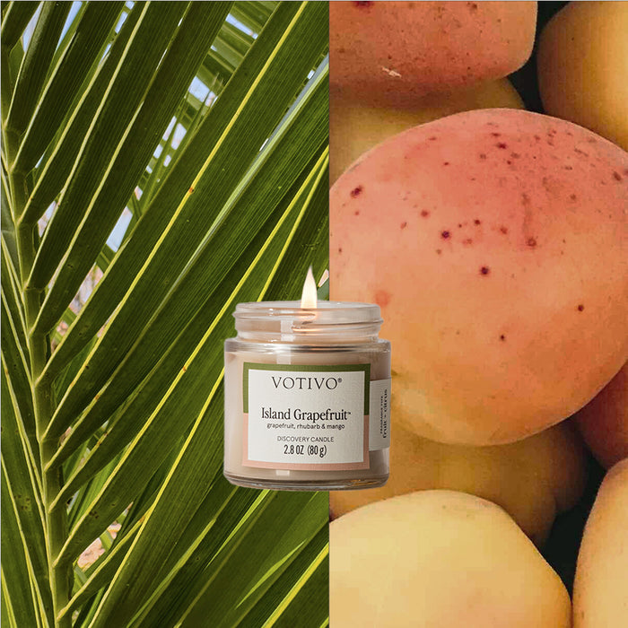 A Votivo Island Grapefruit Discovery Candle with a visible flame glows warmly between close-up green palm leaves and ripe yellow grapefruit. Crafted with a soy blend wax, it adds a vibrant touch to the scene.