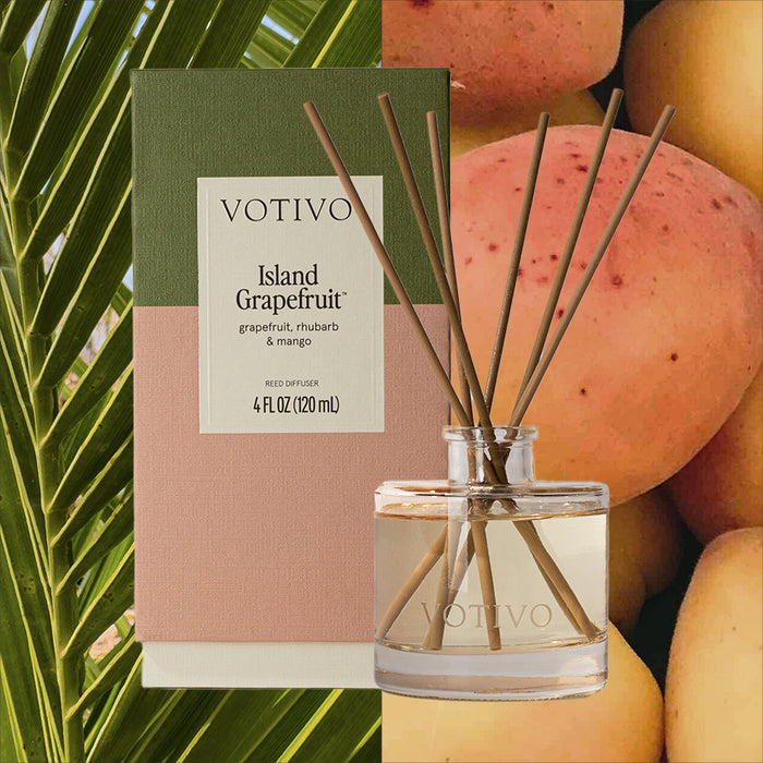 The Votivo Island Grapefruit Reed Diffuser and box are displayed with palm leaves and mangoes; the packaging highlights notes of grapefruit, rhubarb, and mango for a refreshing citrus fragrance.