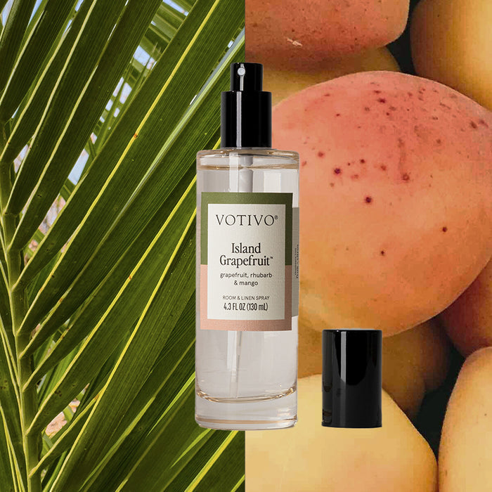 A bottle of Votivo Island Grapefruit Room & Linen Spray with a black cap sits before palm leaves and ripe grapefruit, evoking the vibrant tropical citrus fragrance.