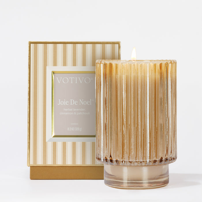 A lit, ribbed glass Joie De Noel Holiday Candle by Votivo sits beside its matching gold and cream striped box, filling the air with herbal lavender, cinnamon, and patchouli notes in a luxurious soy blend wax.