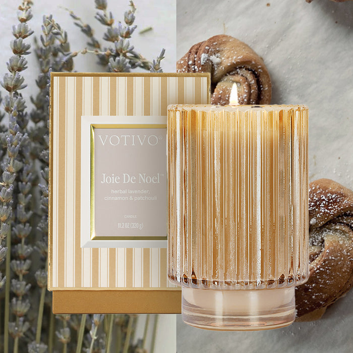 A gold-striped Votivo Joie De Noel Holiday Candle box sits beside a lit ribbed glass Soy Blend Wax candle, with lavender sprigs and powdered sugar-dusted pastries in the background.