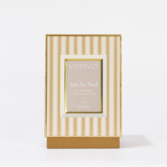 A boxed Votivo Joie De Noel Holiday Candle with herbal lavender, cinnamon, and patchouli scents in a soy blend wax. The packaging features beige and white stripes with gold accents on a plain white background.