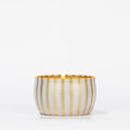 A lit Votivo Joie De Noel Holiday Tin Candle in a round white container with gold stripes, crafted from soy blend wax, is shown against a plain white background.