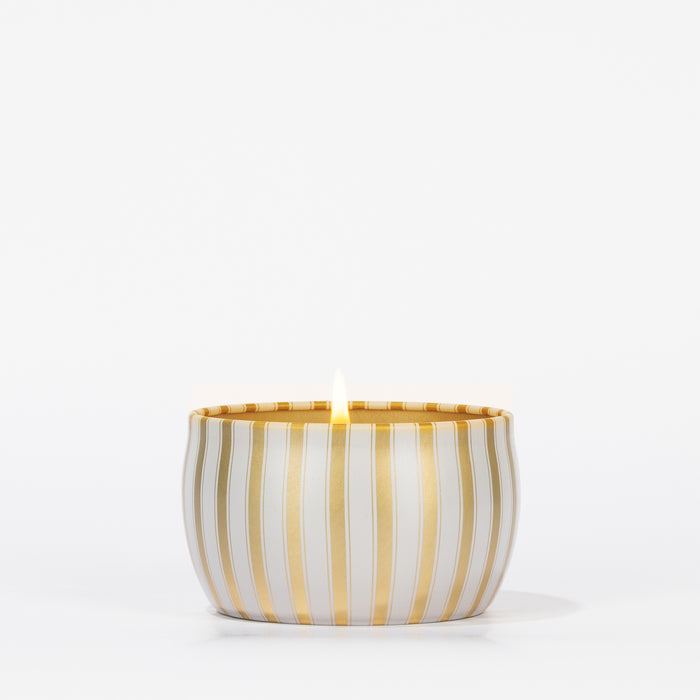 A lit Votivo Joie De Noel Holiday Tin Candle in a round white container with gold stripes, crafted from soy blend wax, is shown against a plain white background.