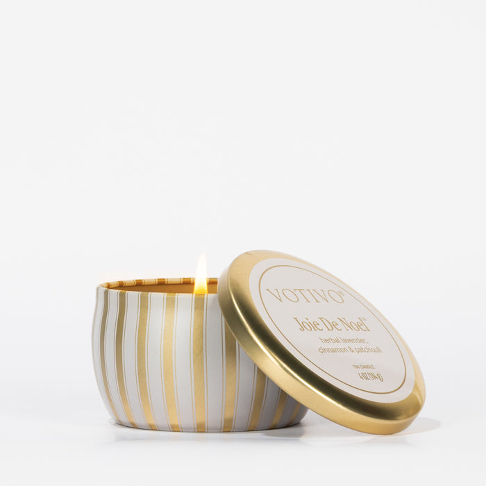 A lit Votivo Joie De Noel Holiday Tin Candle sits in a round tin with a gold and white lid labeled "VOTIVO Joie De Noel." Made with soy blend wax, it is displayed against a plain white background.