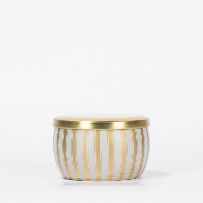 The Votivo Joie De Noel Holiday Tin Candle features vertical gold and white stripes with a matching gold lid, crafted from premium soy blend wax, and is showcased against a plain white background.