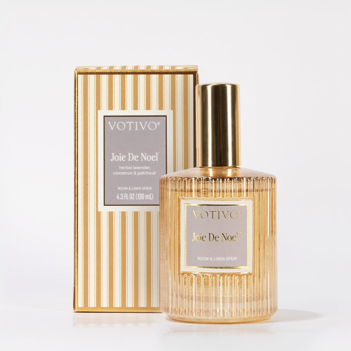 A gold and clear glass bottle of Votivo Joie De Noel Room & Linen Spray is displayed before its matching beige and gold striped box, both bearing the product name—a festive oriental spice fragrance.