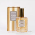 A gold and clear glass bottle of Votivo Joie De Noel Room & Linen Spray is displayed before its matching beige and gold striped box, both bearing the product name—a festive oriental spice fragrance.
