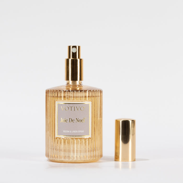 A gold ridged glass spray bottle labeled Votivo Joie De Noel Room & Linen Spray stands upright on a white background, its shiny gold cap placed beside it.