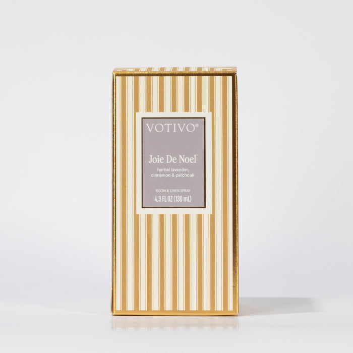 A gold and white vertically striped box of Votivo Joie De Noel Room & Linen Spray, with a gray label and gold border, stands upright on a white background.