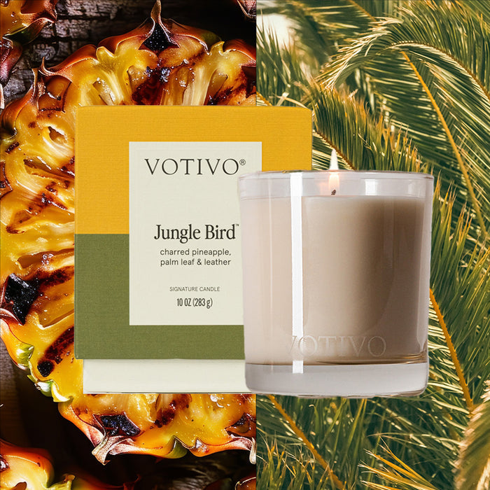 A Votivo Jungle Bird 10 oz. Signature Candle box is placed before a sliced pineapple, beside a lit glass candle crafted with soy blend wax, set against green palm leaves in the background.